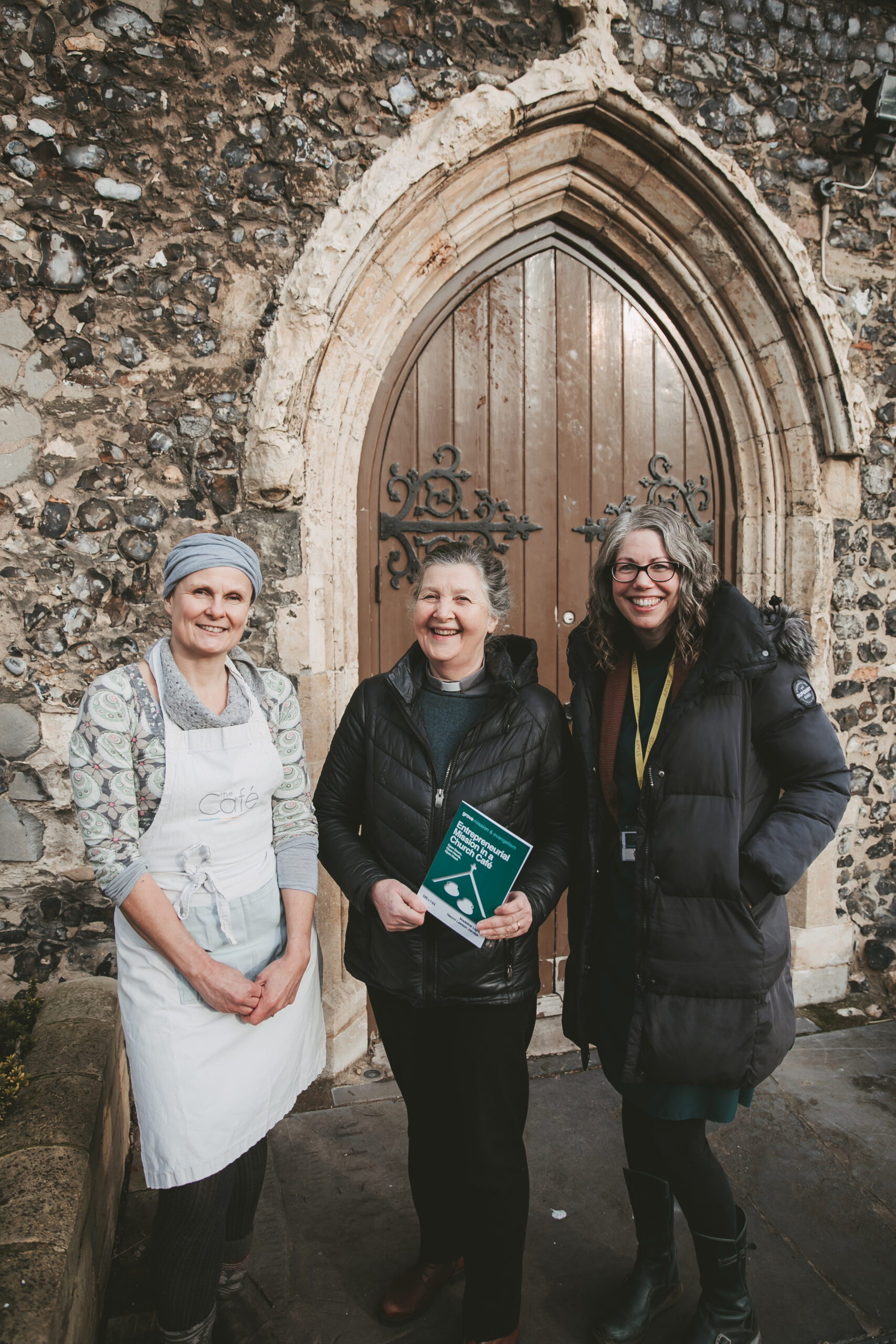 An image of Beckie Ward who is the Cafe manager, the Vicar the Revd Madeline Light and Clare Melia who the Community worker outside an old church door holding a copy of the booklet.